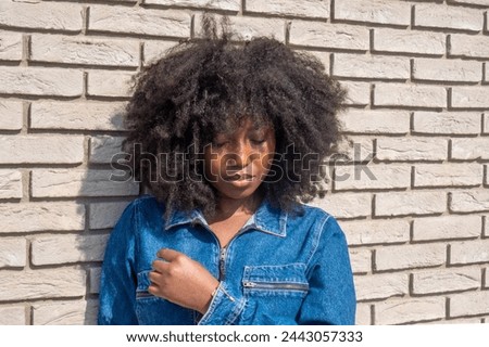 Captured in contemplation, a young African American woman with a full, natural afro stands against a stark white brick wall. The sunlight highlights her denim jacket, adding warmth to the image, while