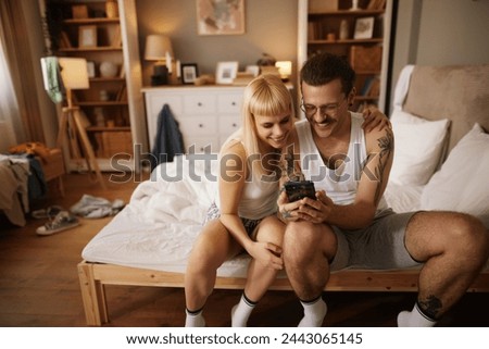 Tattooed man and his wife using smart phone in bed