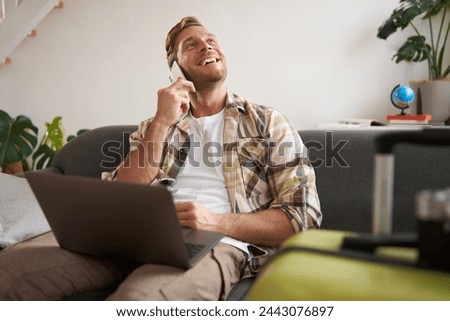 Portrait of young smiling man with suitcase and laptop, making a phone call, booking a flight tickets, calling to confirm hotel room. Copy space Royalty-Free Stock Photo #2443076897