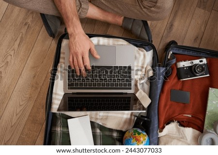 Top view of opened suitcase with clothes, male hands using laptop, booking hotel for vacation, check-in on flight for holiday, packed camera to shoot pictures.