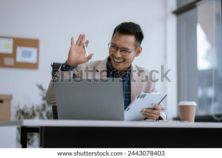 happy smiling Asian male waved to greet colleagues using a smartphone and laptop computer at office. Royalty-Free Stock Photo #2443078403