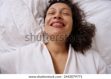 Close-up portrait of a smiling curly young adult woman smiling while waking up in the morning , lying on the bed at home bedchamber. View from above Royalty-Free Stock Photo #2443079771