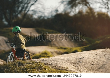 small kid with green helmet rides a balance bike on a dirt jump track in bright sunlight. selective focus on jump in foreground, little child blanked out blurry in background. Family sport concept
