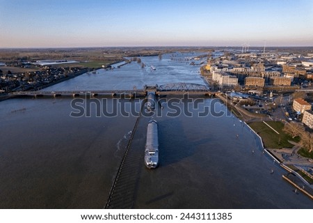 Cargo ships awaiting and passing through steel draw bridge at Zutphen on the overflown river IJssel during high water level. Aerial inland shipping waterway infrastructure concept Royalty-Free Stock Photo #2443111385