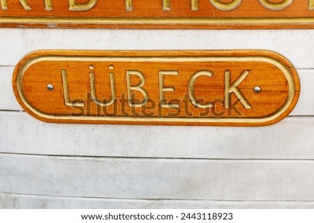 Detailed shot of a nautical nameplate on a boat, with the word LUBECK etched in raised golden letters on a polished wooden background. LÜbeck hanseatic  town sign.