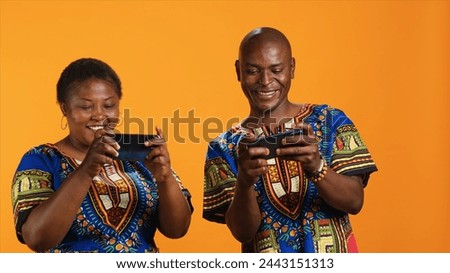 African american couple playing mobile videogames on camera, having fun with online tournament over orange background. Playful happy people enjoying internet contest together.
