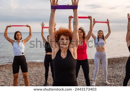 Group of Caucasian adult smiling women in sportswear are training with sports elastic band at coast. Sea on background. Outdoor activity and fitness.