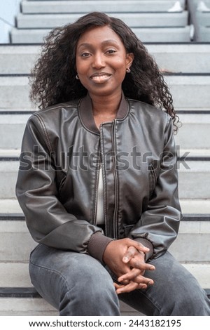 A young African woman with a radiant smile sits comfortably on outdoor steps, her dark curly hair framing her face. She wears a fashionable leather jacket, presenting a relaxed yet confident posture Royalty-Free Stock Photo #2443182195