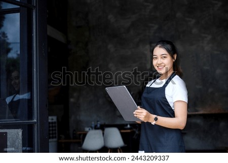 Pretty Asian coffee shop owner  in white t shirt and apron standing in front of shop while holding tablet and smiling to the camera