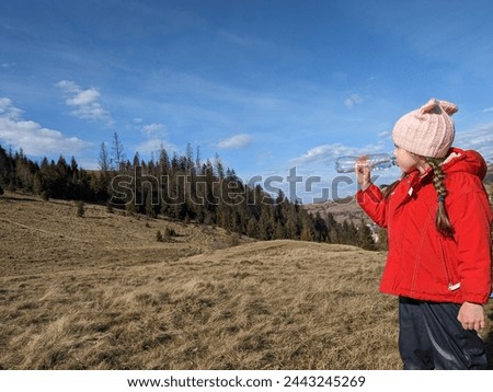 
A girl drinks water from a plastic bottle on a mountain slope against the backdrop of a spruce forest, active family vacation. Photo.