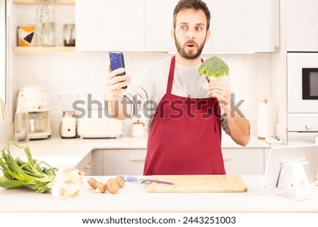 A man is taking a picture of a broccoli in his kitchen. He is wearing a red apron and standing in front of a counter with various kitchen appliances such as a toaster, oven, and microwave