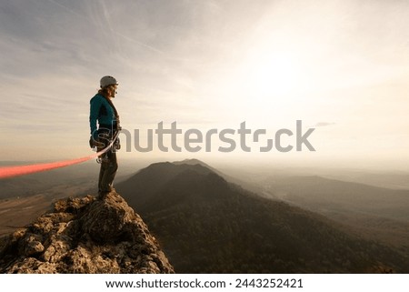 A woman is standing on a mountain top with a red rope tied to her. The sky is cloudy and the sun is setting