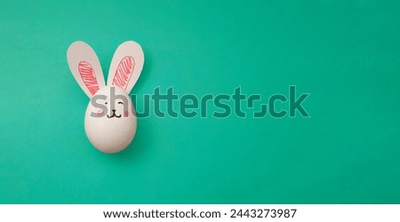 White chicken egg with pink rabbit paper ears on green background. Horizontal banner with Easter Bunny. Copyspace for text. Hare muzzle. Preparations for celebration of religious holiday Happy Easter