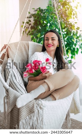 Vertical image of cute smiling brunette woman with bunch of flowers in hands is relaxing and swinging on hanging chair. Present in women's day from beloved man. St. Valentines day