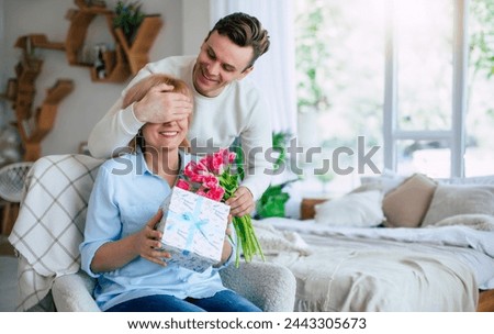 Close up image of young smiling man while he making surprise for his mature mother with some holiday in domestic room. Mothers day, birthday or womens day concept