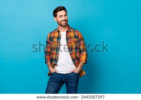 Portrait of optimistic guy with stubble wear checkered shirt arms in pockets look at offer empty space isolated on blue color background