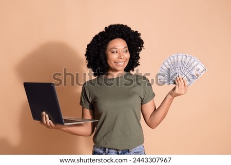 Portrait of satisfied girl dressed t-shirt hold laptop look at dollars in arm get impressive income isolated on beige color background