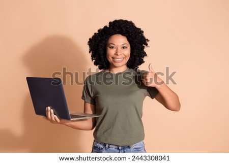 Portrait of multiethnic multinational girl dressed t-shirt hold laptop showing thumb up nice job isolated on beige color background Royalty-Free Stock Photo #2443308041