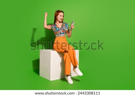Full size photo of pretty young woman sit cube raise fists hold phone wear top isolated on bright green color background