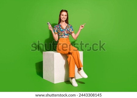 Full size photo of pretty young woman sit cube hold phone point finger empty space wear top isolated on bright green color background