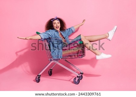 Full length photo of cheerful carefree lady dressed denim outfit arms sides riding shopping cart empty space isolated pink color background