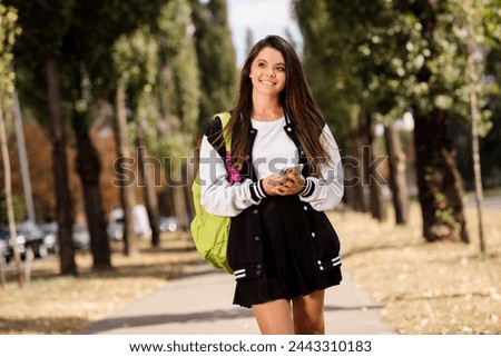 Photo of cheerful student with straight hairdo dressed stylish jacket rucksack on shoulder hold smartphone enjoy sunny weather outdoors