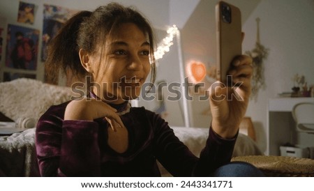 Beautiful teenager sits on floor near bed in her room and takes a pictures using phone. African American girl chats with friends or creates blog content. Spending leisure time at comfortable home.