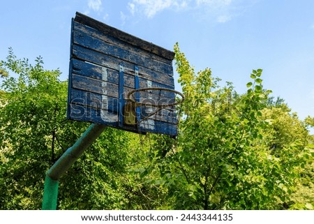 A basketball hoop with a blue backboard and a rusty net. The net is hanging loose and the backboard is missing a piece. The hoop is located in a wooded area with trees surrounding it