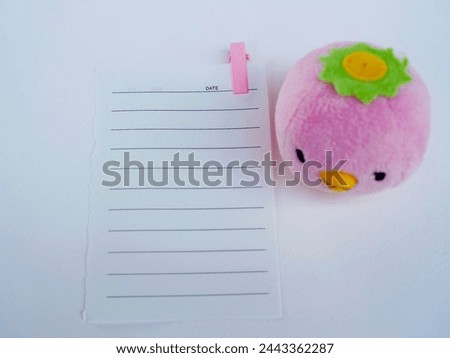 A blank piece of paper for writing on has a pink clip attached to it and a pink baby bird doll sits on a white background.