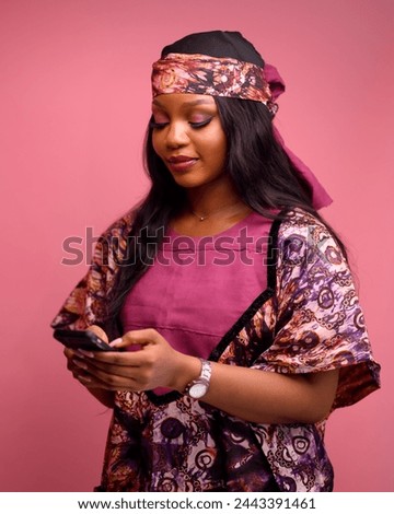 Friendly young stylish African-American woman chatting and looking at her phone. A confident successful dark-skinned woman in fashionable peach native attire copy space pink background	