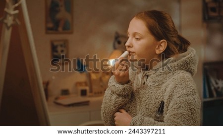 Young beautiful girl does makeup standing and taking picture in the mirror in cozy bedroom using mobile phone. Caucasian teenager spending leisure time at home in the evening. Concept of lifestyle.