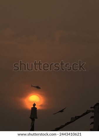 As we can see in this picture the sun is about to set and the helicopter is flying over from top and we can also flying bird in this picture. Beautiful picture and good timing to capture this picture.