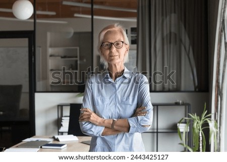 Serious confident blonde senior business woman in elegant glasses looking at camera with arms folded, standing at modern office workplace. Mature manager, director posing for professional portrait