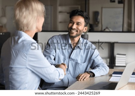 Happy young Indian entrepreneur man in casual shaking hands with senior professional colleague woman at office workplace table, smiling, thanking for support, enjoying business communication
