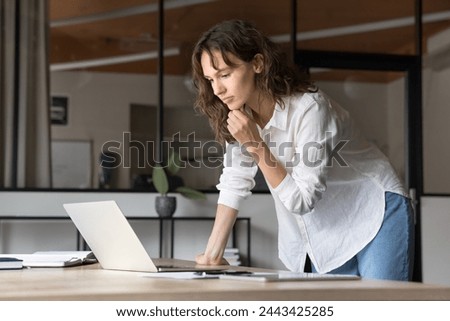 Thoughtful engaged young manager woman using laptop at workplace for online job communication, working at computer in office, standing at table, touching chin, looking at computer, thinking