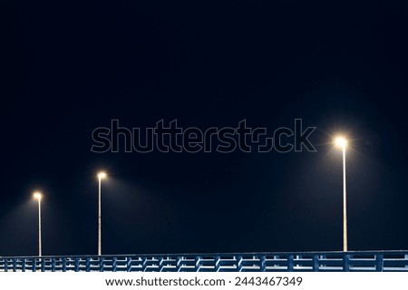 Street lampposts casts dim cold blue glow along shore pier standing against pitch black canvas of night sky paints poetic picture of pier in stillness of night, serenity of solitary moments