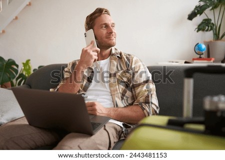 Handsome smiling man with laptop, making a phone call, looking aside while waiting on the telephone, confirms booking online. Tourism concept Royalty-Free Stock Photo #2443481153