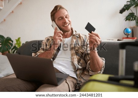 Portrait of young man with credit card, sitting and using laptop, making a phone call, booking hotel or tickets, confirming his purchase over telephone, has suitcase near him. Royalty-Free Stock Photo #2443481201
