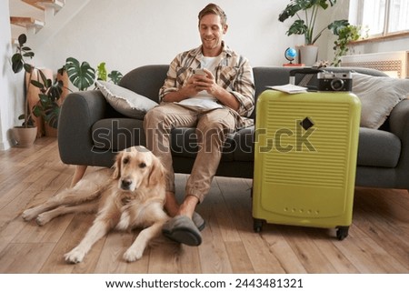 Portrait of guy with a dog, has packed suitcase, camera for going abroad and shooting pictures, dog resting near his legs, man looks at mobile phone and chats online.