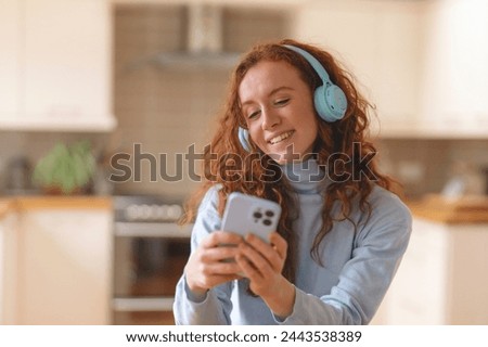Happy young redhead woman in blue shirt using a mobile phone, listening to favorite songs, audiobooks, podcasts on headphones while sitting in kitchen at home