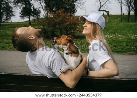 Young Caucasian couple is sitting on wooden bench with a jack Russell terrier puppy on spring sunny day. People with a dog in Kalemegdan Park in Belgrade. Dog wants to bite playfully and shows teeth