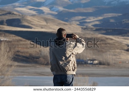 A man in a hooded jacket takes pictures of mountains and a lake on his phone