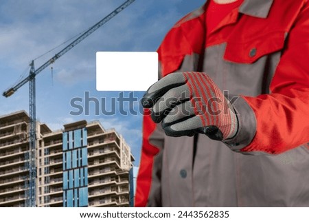 A man in work clothes and gloves holds a blank plastic card in his hands. Against the backdrop of the construction of a new house.