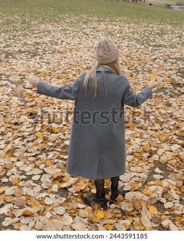 A young woman in a gray coat among autumn leaves in a city park. Photograph with a predominantly yellow color due to the large number of leaves of that color in the ground.