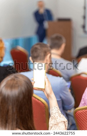 A woman shoots a lecture on a smartphone camera in a business conference or seminar. Audience listens to the lecturer at the conference in auditorium.
