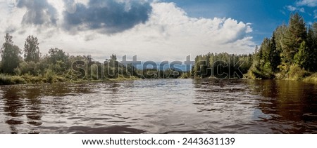 A wide river gently flows through a lush forest under a sky dotted with billowy clouds Royalty-Free Stock Photo #2443631139