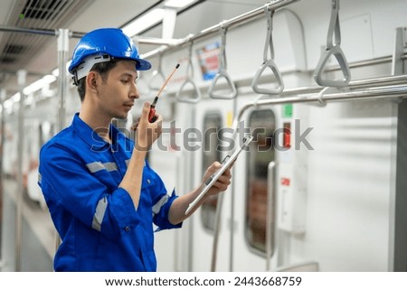 A young train maintenance engineer is using a radio to communicate while holding a clipboard, with a colleague in the background.

