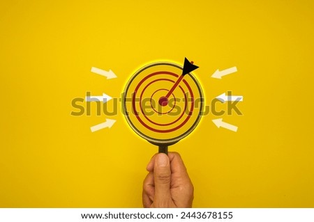 A person is holding a magnifying glass over a yellow background with a red arrow pointing to the center. Concept of focusing on a specific target or goal