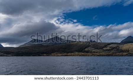 The beautiful landscape of Patagonia. The Andes mountains on a background of blue sky and clouds. The sparse vegetation of the coastal strip. There are ripples on the blue water of the Beagle Canal.