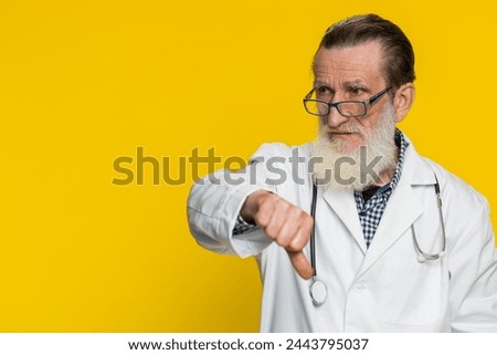 Dislike. Upset unhappy senior doctor cardiologist man showing thumbs down sign gesture disapproval dissatisfied negative feedback. Apothecary pharmacy grandfather on yellow background. Copy-space